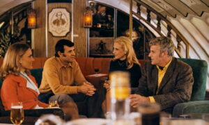 Four adults sit on a retro-style sofa in an airplane lounge, chatting over drinks in a warmly lit cabin interior.