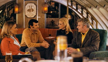 Four adults sit on a retro-style sofa in an airplane lounge, chatting over drinks in a warmly lit cabin interior.