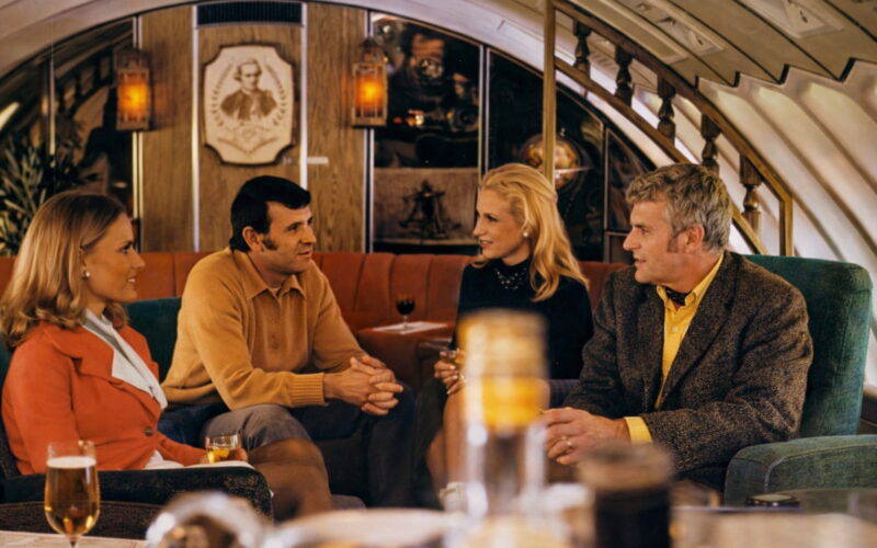 Four adults sit on a retro-style sofa in an airplane lounge, chatting over drinks in a warmly lit cabin interior.