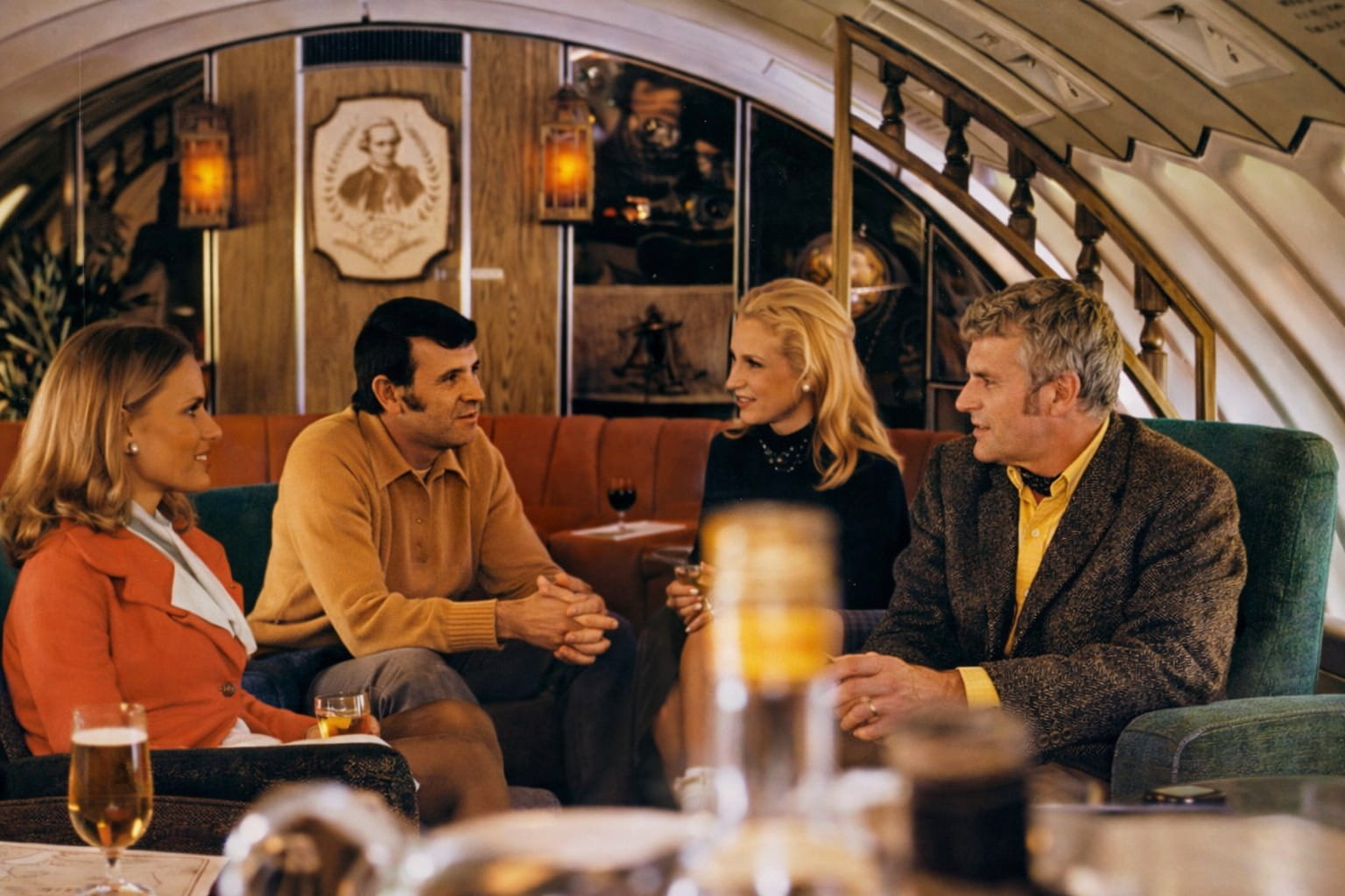 Four adults sit on a retro-style sofa in an airplane lounge, chatting over drinks in a warmly lit cabin interior.