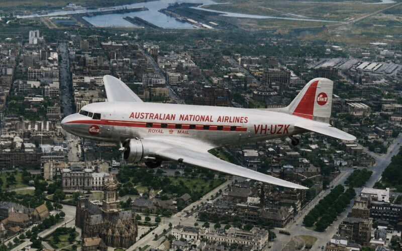 Vintage silver airliner with red stripe reading 'AUSTRALIAN NATIONAL AIRLINES' over a city, registration VH-UZK, early jet era styling with a cathedral visible below.