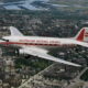 Vintage silver airliner with red stripe reading 'AUSTRALIAN NATIONAL AIRLINES' over a city, registration VH-UZK, early jet era styling with a cathedral visible below.