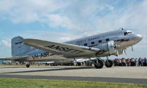 Silver vintage propeller airliner with ANSETT markings on the fuselage, taxiing on a runway as a crowd watches.