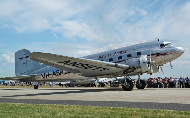 Silver vintage propeller airliner with ANSETT markings on the fuselage, taxiing on a runway as a crowd watches.