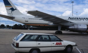 Large white airliner on the tarmac with a white sedan marked 'AWA' in the foreground.