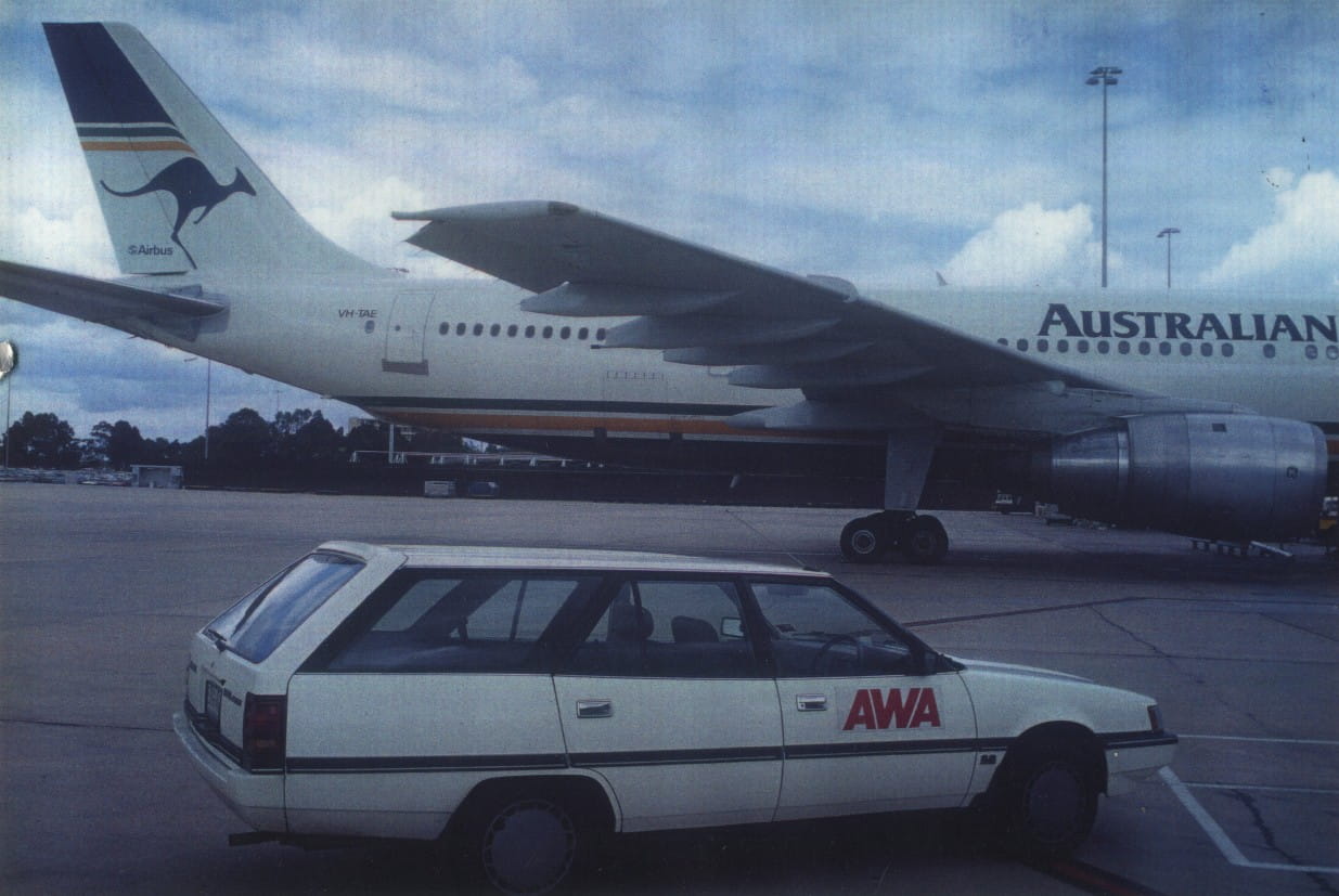 Airbus airliner at the airport with a kangaroo logo on the tail; a white AWA station wagon is parked in the foreground.